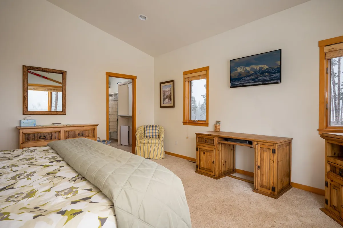 A bedroom featuring a bed with floral bedding, wooden furniture, a mirror, wall artwork, and a cozy desk area. Natural light pours in through the windows, illuminating the tranquil space ideal for restful nights and peaceful mornings.