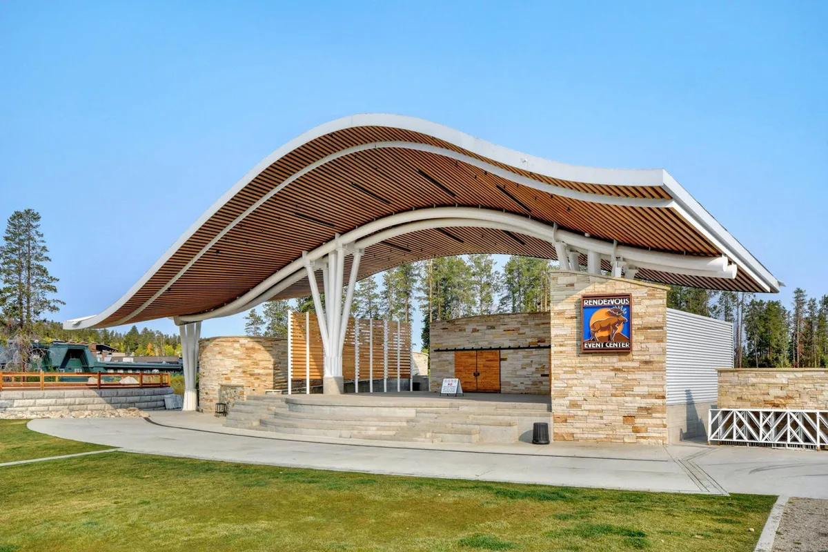 An exterior shot of a community event space with a curved roof structure, sturdy stone walls, and warm wooden accents. The scene suggests a vibrant, festive atmosphere suited for live music and food festivals.
