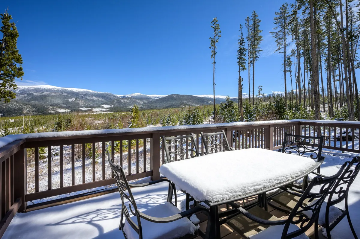 A serene outdoor setting featuring a snow-covered table with a panoramic mountain view and tall trees surrounding the patio. The sun is setting, casting a warm glow over the landscape, evoking a tranquil and private alpine retreat atmosphere.