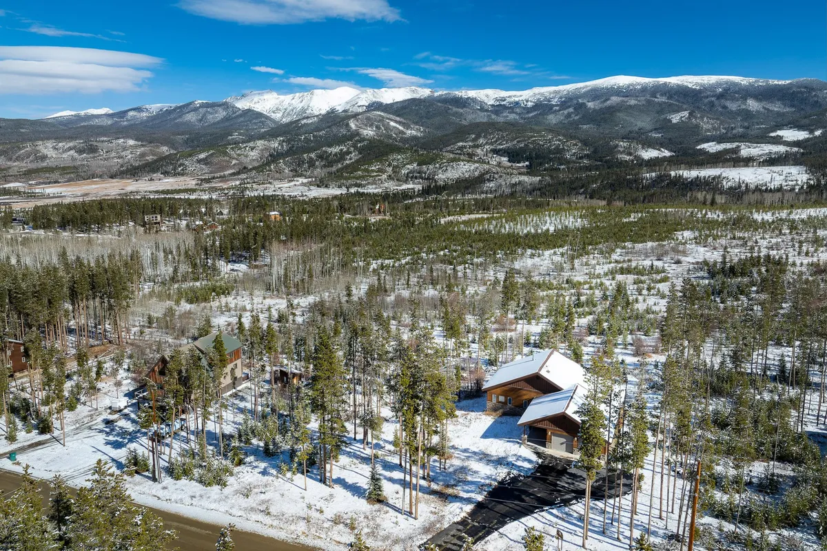 A wide exterior scene featuring snow-covered mountains, tall pine trees, and cozy cabins nestled beneath a clear blue sky. The landscape transitions easily between seasons, with the setting evoking both winter sports and summer hiking adventures.