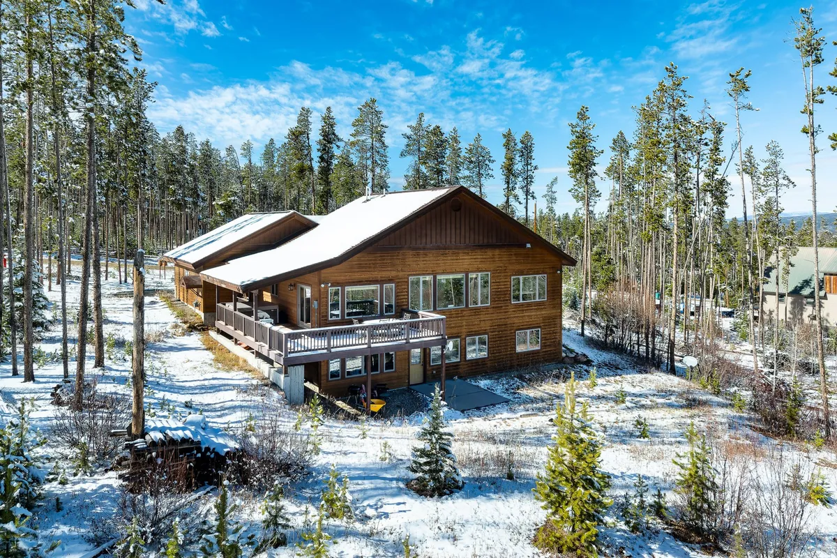 The image showcases a spacious wooden mountain cabin nestled among snow-covered pine trees in Fraser, Colorado. The scene highlights the cabin's large deck, perfect for enjoying the breathtaking alpine views, and evokes a sense of warmth and coziness amid the wintry landscape.
