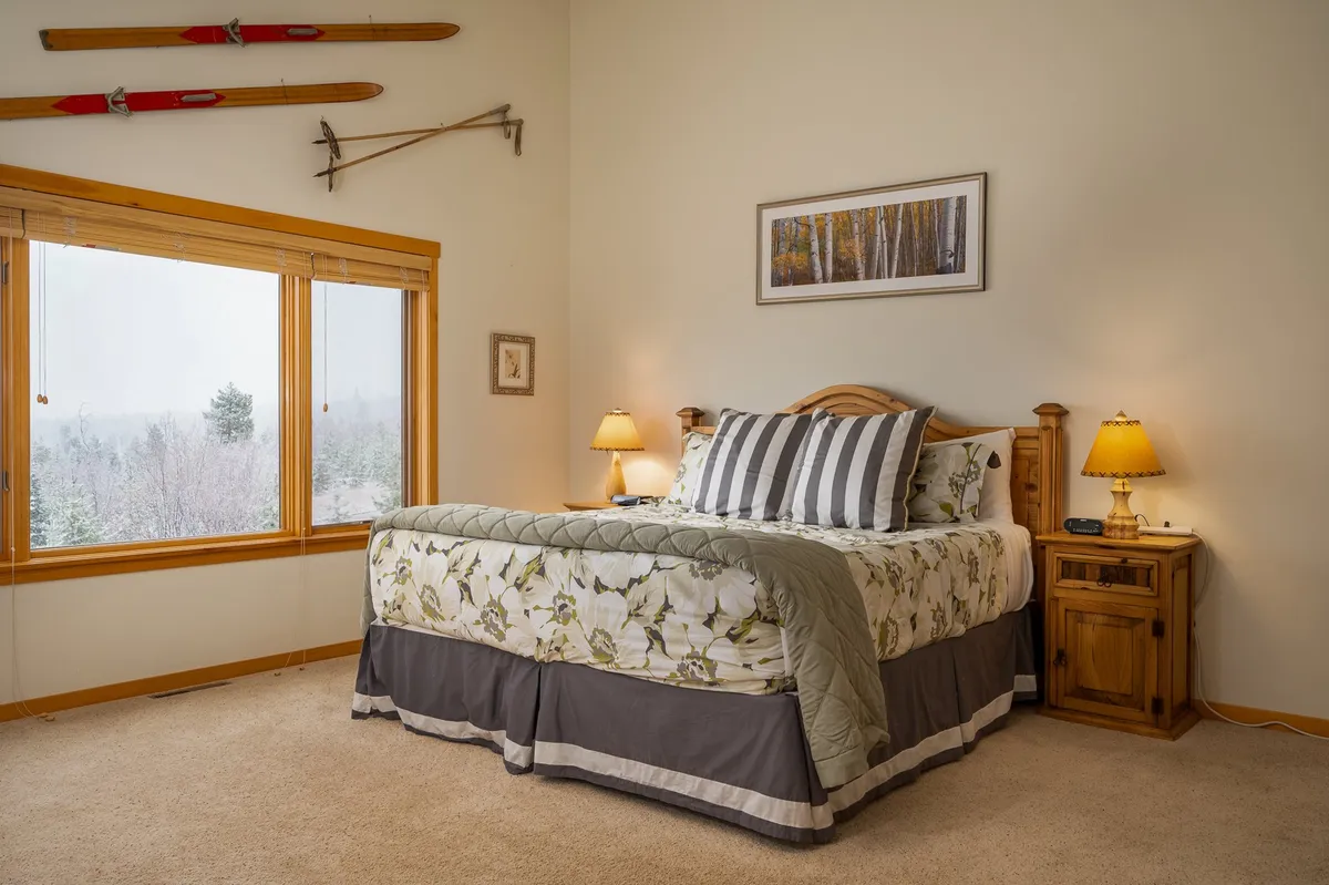 A tranquil bedroom with a comfortable bed, large window overlooking nature, and ski equipment neatly stored. Nightstands flank the bed, and morning sunlight streams in, offering a peaceful retreat with beautiful Colorado views.