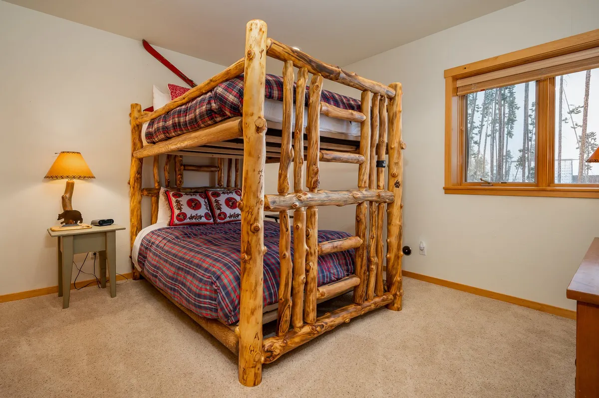 A log bunk bed with plaid bedding sits beside a window offering a glimpse of the outdoors. A nightstand with a lamp provides a warm, family-friendly ambiance, perfect for both children and adults to relax and unwind.