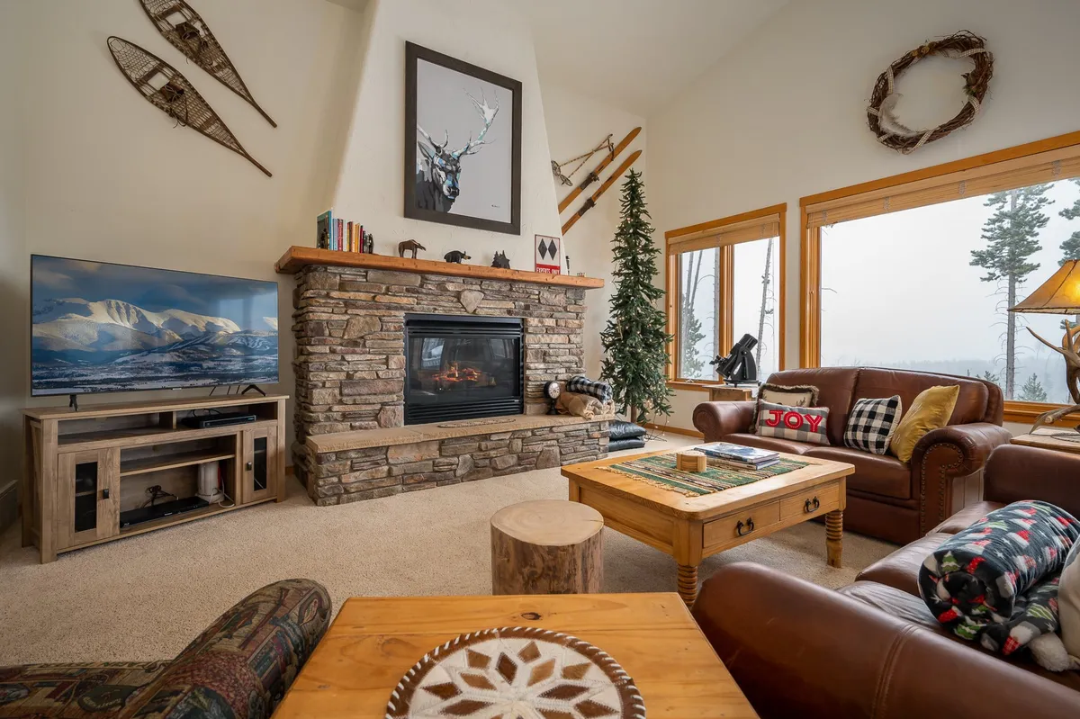 A spacious living room featuring a striking stone fireplace, large windows that reveal panoramic mountain views, a comfortable leather sofa, a wooden coffee table, and tasteful decorative accents. The setup is inviting, perfect for gathering, watching TV, or relaxing with family.