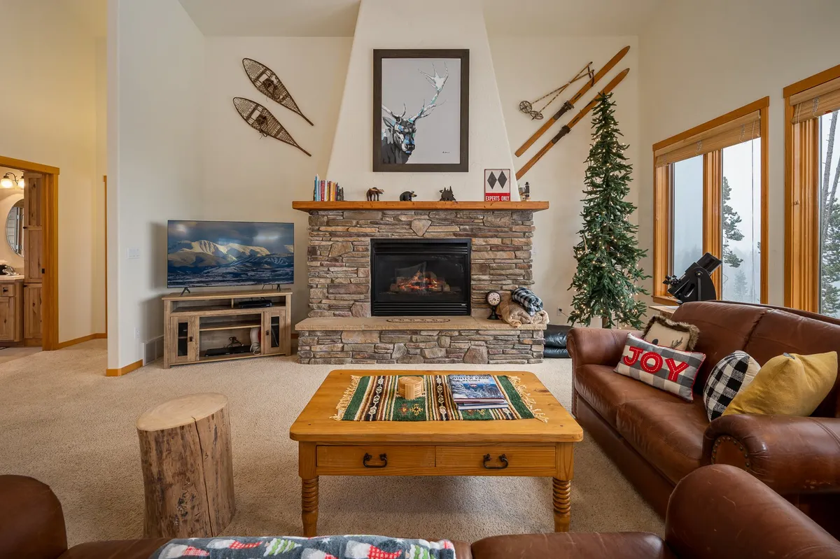 A cozy living room with a stone fireplace, leather sofas, a wooden coffee table, decorative deer artwork, and a tall Christmas tree. The space is perfect for indoor entertainment—hosting game nights, gathering for movies on the smart TV, or simply unwinding by the fire.