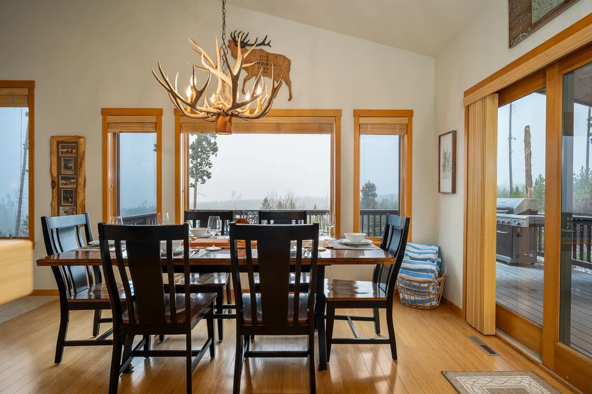 A large dining area centered around an expansive table set for 12, accented by a stylish chandelier overhead. The space is surrounded by tall windows showcasing scenic views and decorated with art, creating an atmosphere ideal for shared meals and celebrations.