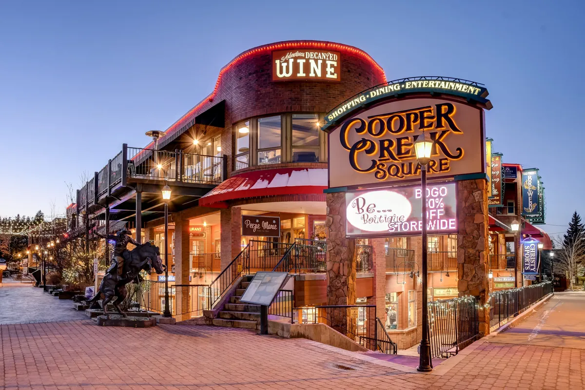 A bustling downtown area with the Cooper Creek Square sign, inviting wine shop, outdoor seating, and festive string lighting, suggesting a lively hub for shopping, dining, and socializing.